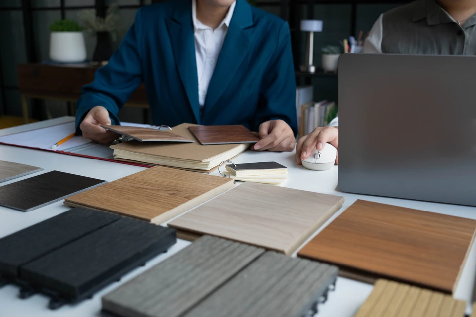Homme d'affaires souriant en cardigan gris et chemise blanche présentant des documents contractuels à une cliente blonde lors d'une consultation professionnelle dans un bureau moderne avec baies vitrées