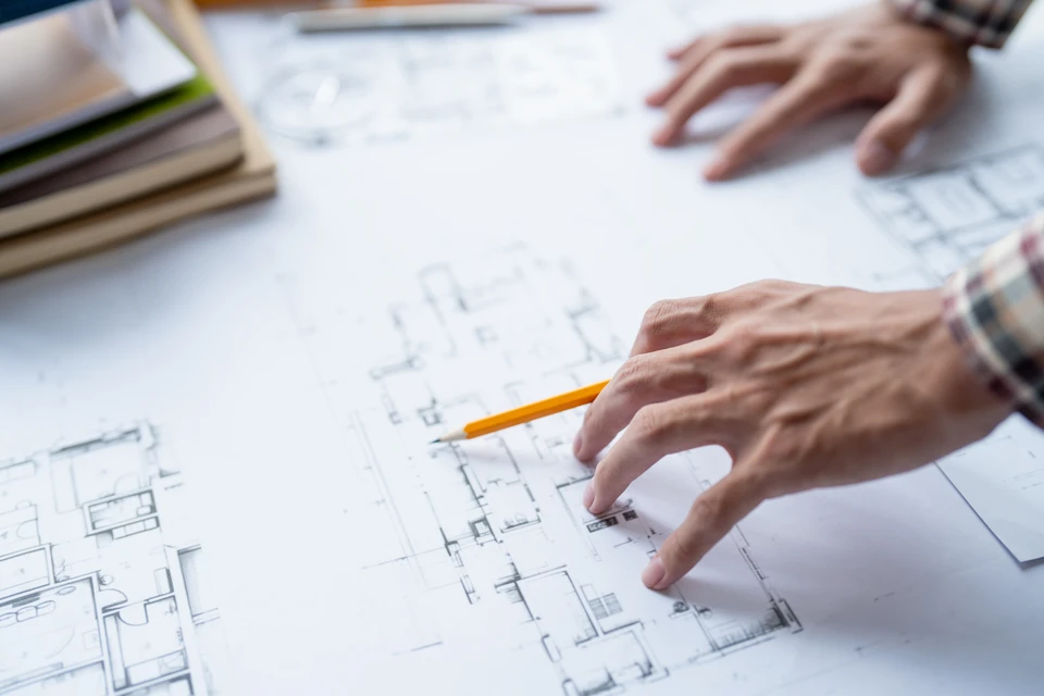 Un chef de projet ou manager de chantier en tenue professionnelle d'entreprise de b&acirc;timent (chemise, gilet de s&eacute;curit&eacute; ou costume), consulter des plans de r&eacute;novation sur un chantier multi-corps de m&eacute;tier, avec visible en arri&egrave;re-plan des artisans et ouvriers travaillant &agrave; diff&eacute;rentes t&acirc;ches (menuiserie, plomberie, &eacute;lectricit&eacute;), symbolisant la coordination et l'orchestration de plusieurs m&eacute;tiers du b&acirc;timent pour une r&eacute;novation globale.